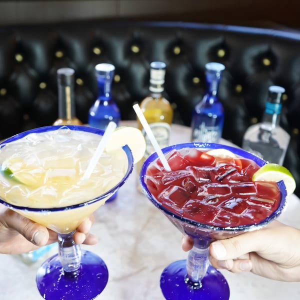 Two hands holding blue-rimmed margarita glasses, one yellow and one red, with tequila bottles behind.