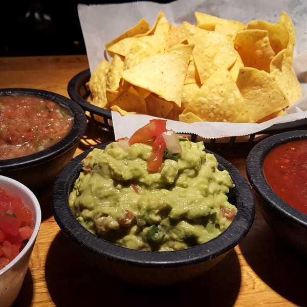 A basket of tortilla chips with bowls of guacamole, salsa, and diced tomatoes on a wooden table.